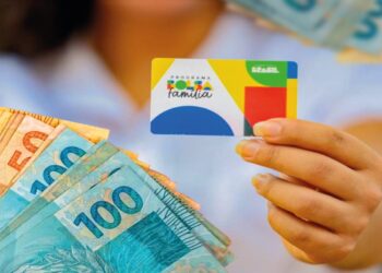 Pessoa segurando o cartão do Bolsa Família com dinheiro em notas ao fundo