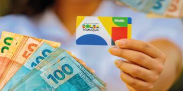 Pessoa segurando o cartão do Bolsa Família com dinheiro em notas ao fundo